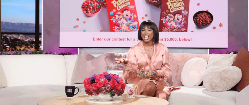 Jennifer Hudson sits on a couch with Fruity and Cocoa PEBBLES Crunch'd on a background screen.