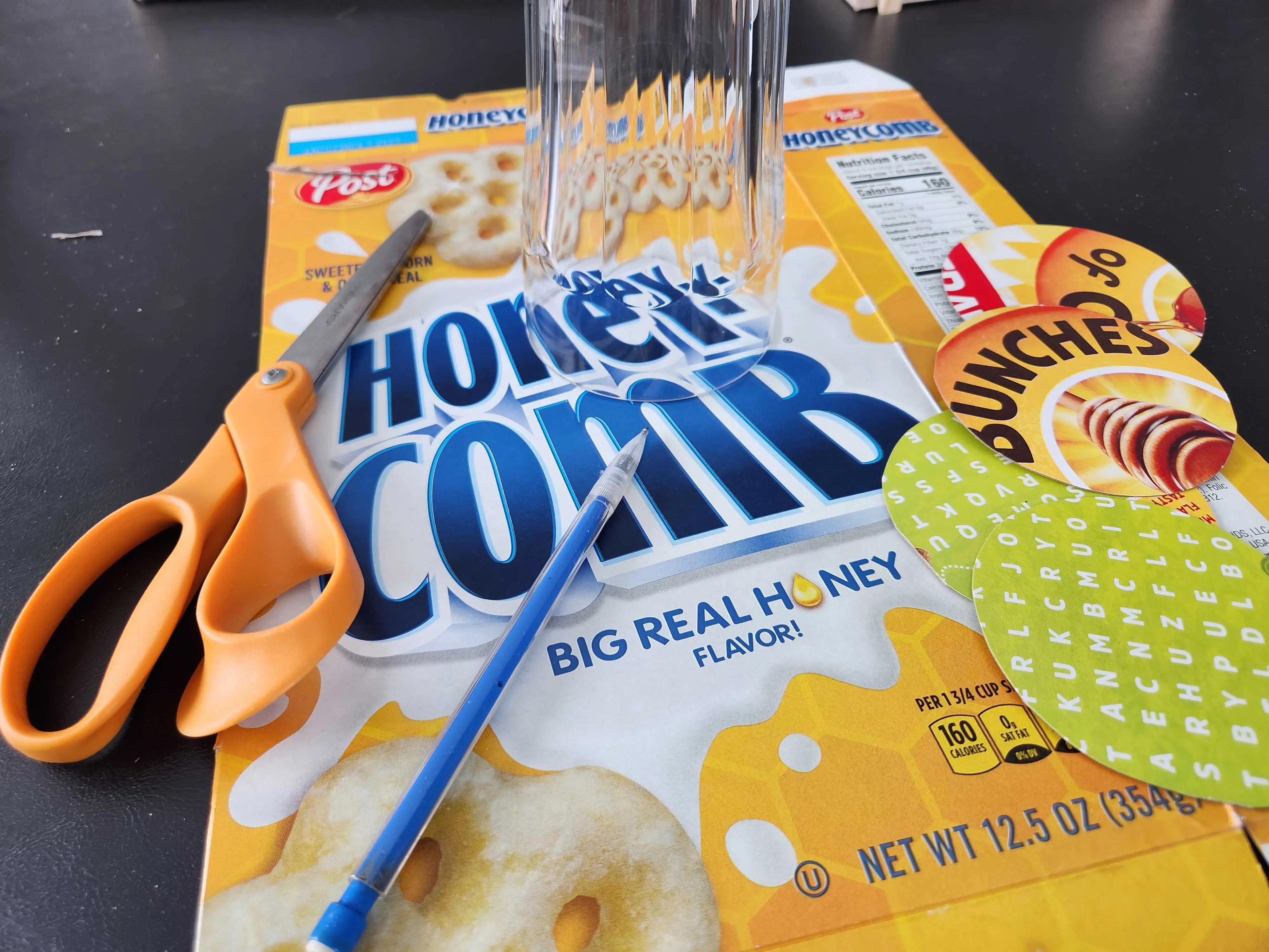 A close-up of a box of Honeycomb cereal with scissors, a glass and a pen on it.