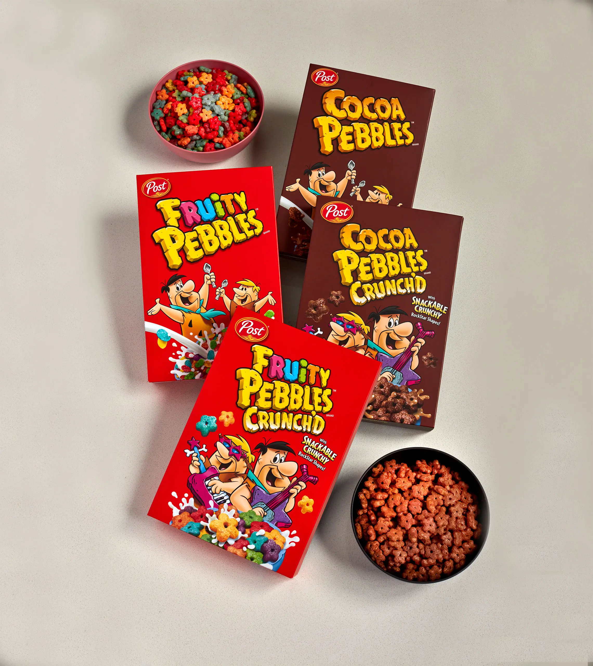 A top down view of boxes of FRUITY PEBBLES and COCOA PEBBLES CRUNCH'D. There are bowls of the cereals on the top left and bottom right.