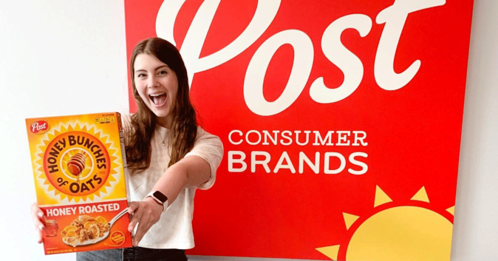 Summer intern holding Honey Bunches of Oats cereal box in front of Post Consumer Brands sign